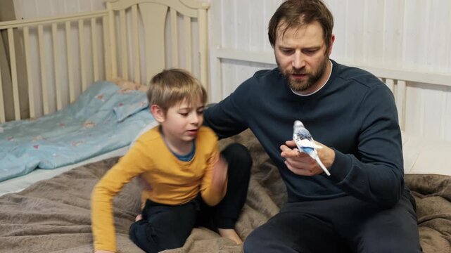 Father and son enjoying at home. Young boy and his daddy playing with funny pet budgerigar parrot. Happy family. Tamed budgie parakeet. People take care of and play with bird. Cute domestic animals