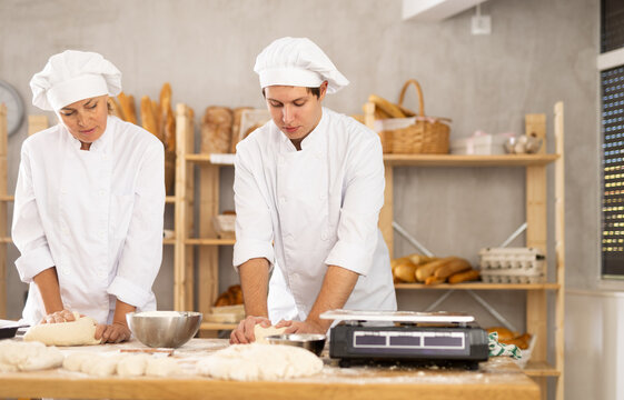 Adult woman and young man bakers kneading dough in bakery workshop