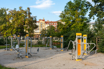 Outdoor fitness equipment with yellow and gray exercise machines in public park surrounded by green trees and residential buildings. Healthy lifestyle promotion and community wellness facilities.