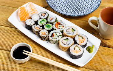 Hearty lunch has been prepared - uramaki sushi set and nigiri with avocado, cucumber, soft cheese and salmon on white plate, supplemented with bowl of soy sauce and chopsticks