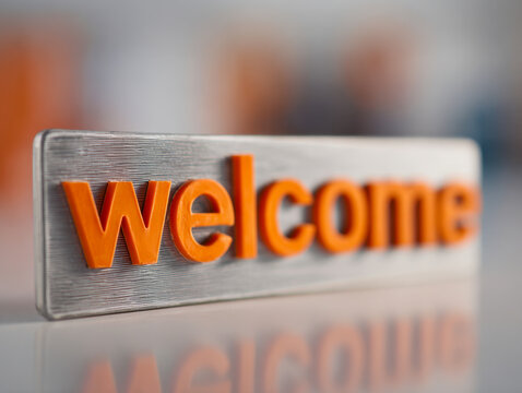 Modern metallic sign with embossed bold orange text spelling out a warm greeting indoors with a clean reflective surface and blurred background in soft focus