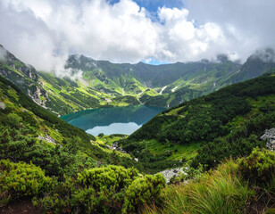 Mountain lake surrounded by green hills and cloudy sky, peaceful nature landscape with fresh vegetation and rocky terrain under soft sunlight