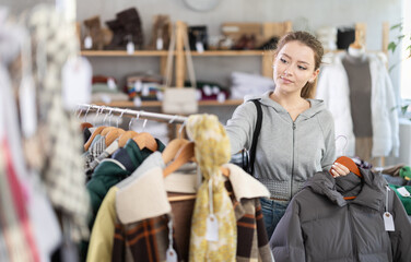 Young woman with interest chooses a stylish and warm down jacket for autumn and winter in a clothing store. Customer stands among the shelves and hangers in the store © JackF