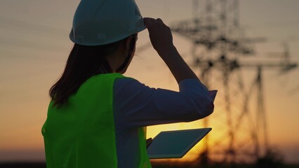 Woman engineer, power engineer in helmet checks power line using computer tablet online. Electrician in outdoors. Electric lines of high voltage at sunset. Distribution and supply of electricity