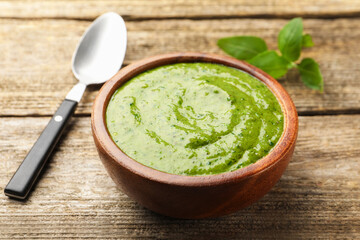 Tasty green pesto in bowl and basil leaves on wooden table, closeup