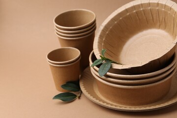 Set of disposable tableware and green leaves on brown background, closeup. Space for text