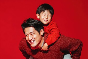 A joyful Asian father carries his son on his back against a red backdrop enjoying indoor playtime together