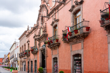Obraz premium A row of buildings with balconies and potted plants. The buildings are pink and the sky is cloudy. Historic center of Querétaro, colonial architecture, decorations for the celebration of Mexico's Inde