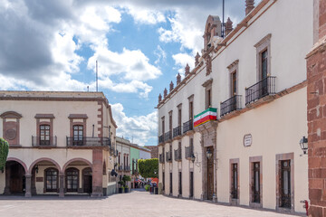 A small town square with a white building in the background. The building has a flag on it. Historic center of Querétaro, colonial architecture, decorations for the celebration of Mexico's Independenc