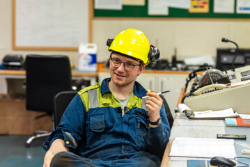 Male engineer in a blue and green coverall with a yellow safety helmet and glasses, sits in the...