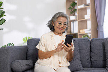 Obraz premium Asian Elderly woman sitting on gray sofa smiling while using smartphone in cozy living room with plants and bookshelves, enjoying relaxed moment at home
