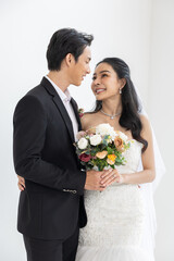 Asian happy bride and groom looking at each other lovingly while holding colorful bouquet of flowers, dressed in elegant wedding attire for their special day