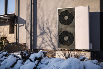 Modern air source heat pump installed outside a residential building in winter. Snow-covered plants and sunlight reflect the concept of sustainable home heating and energy efficiency.