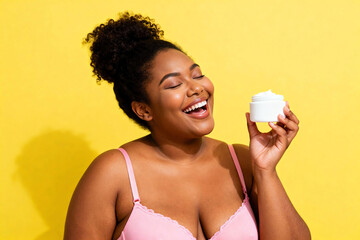 Joyful plus-size Black woman laughing with eyes closed, holding a white skincare cream jar on a vibrant yellow background, celebrating beauty and self-care.