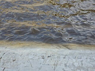 Close-up showing where rippling, murky floodwater on the road meets the rough, horizontal edge of light gray concrete pavement after heavy rain.