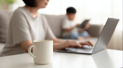 Woman working on laptop with coffee cup in foreground and child using tablet on sofa in background