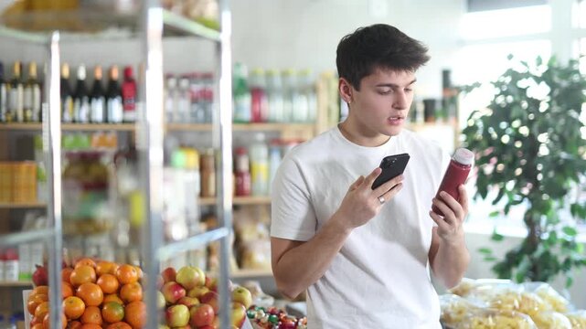 Focused interested young guy scanning barcode on colorful bottles with fresh fruit fruit smoothie using phone while choosing vitamin drinks in grocery store, checking product information and price