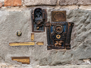 Artistic detail of the smallest clock on the wall near the Leaning Clock Tower in Tbilisi, Georgia, featuring brass plaques, symbolic engravings, and creative steampunk-style street installation.