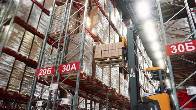 A worker on a forklift lifts boxes of goods and places them on a shelf of a rack in a large modern distribution center. Warehouse filled with cardboard boxes on shelves A shipping and supply chain
