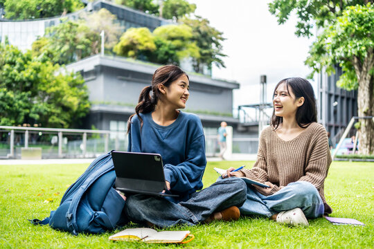 Two young asian female college friends are sitting on the grass at university, happily tutoring and use tablet computer each other. Their smile and laughter show the joy of learning together