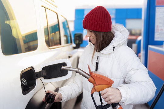 A woman in a white down jacket refueling her car at a gas station. She holds a gun in her hands to fill the car's gas tank. - Powered by Adobe