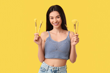 Beautiful young woman with makeup brushes on yellow background