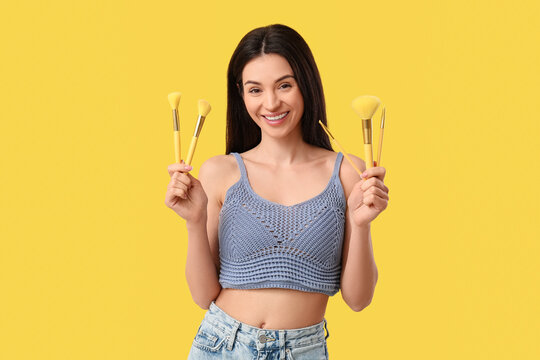 Fototapeta Beautiful young woman with makeup brushes on yellow background