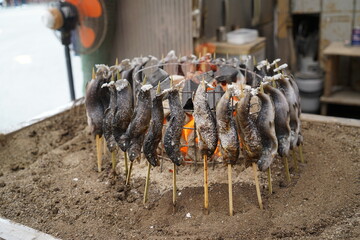 Traditional Japanese style fish grilling with skewers arranged in a circle over charcoal fire, outdoor street food preparation showcasing rustic cooking method and authentic culinary culture.