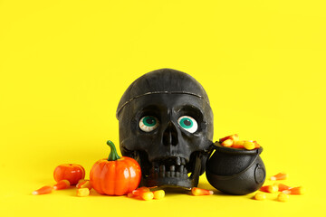 Cauldron with tasty treats for Halloween, pumpkins and skull on yellow background, closeup