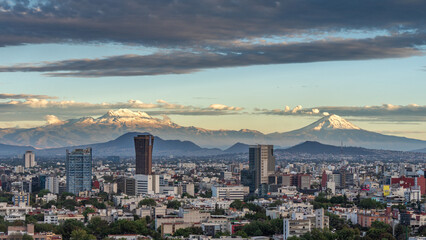 Obraz premium A city with a mountain range in the background. The sky is cloudy and the sun is setting. Panoramic view of Mexico City, snow-covered volcanoes Popocatepetl and Iztaccihuatl