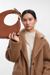asian woman wearing a beige coat holds a large circular leather bag in a bright studio, presenting a calm chic mood with clean lighting and a minimalist fashion vibe, creating a refined and