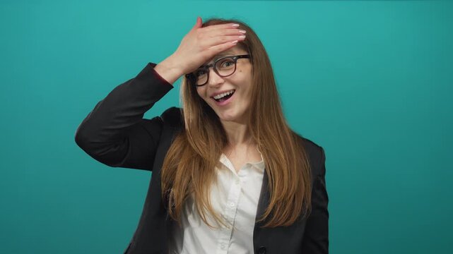 Woman wearing glasses and black blazer over white shirt with long hair smiles and touches forehead in studio; pleasant surprise.