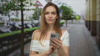 Young caucasian woman holds smartphone with both hands and scrolls screen on busy city street; digital connection. - Powered by Adobe