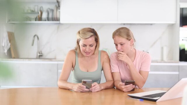 Positive adult woman with teen daughter engrossed in phones sitting at table in cozy home kitchen. Concept of family phubbing and digital distractions