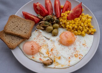 Healthy breakfast with fried eggs, corn, olives, mussels and fresh tomatoes served with whole grain toast on a plate. Nutritious protein meal, balanced diet food, Mediterranean breakfast close-up.