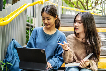 Two beautiful young Asian friends sitting on the stairs at university school, happily tutoring and use tablet computer with book each other. Their smile and laughter show the joy of learning together