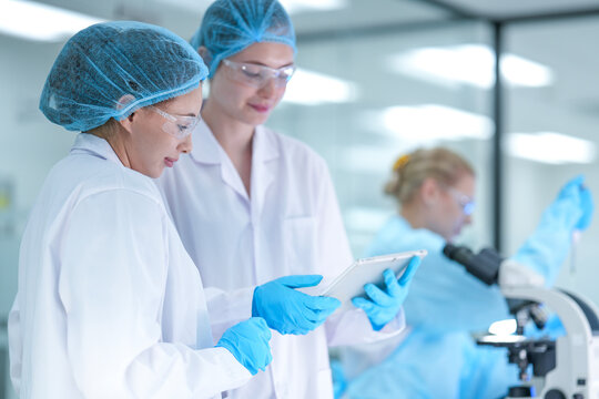 A senior scientist mentors a junior researcher, pointing at results on a digital tablet as they collaborate on an experiment in a bustling, modern scientific laboratory environment.