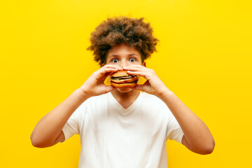 surprised curly african american boy eating burger on yellow isolated background, shocked hungry...