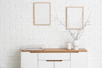 Vase with tree branches on chest of drawers and blank frames near white brick wall