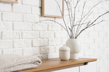 Vase with tree branches on chest of drawers near white brick wall