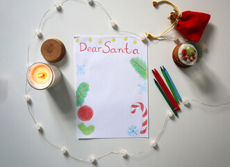 Flat lay of a Dear Santa letter with Christmas decorations, burning candle, red pouch, garland, crayons and snow globe on white table. Letter template drawing by colored pencils. Children wish list.