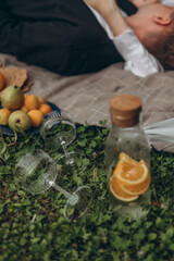 Summer picnic composition with jug of water and orange slices, empty glasses and plate of fruits on brown blanket. Romantic couple lying on grass, bodies partly visible