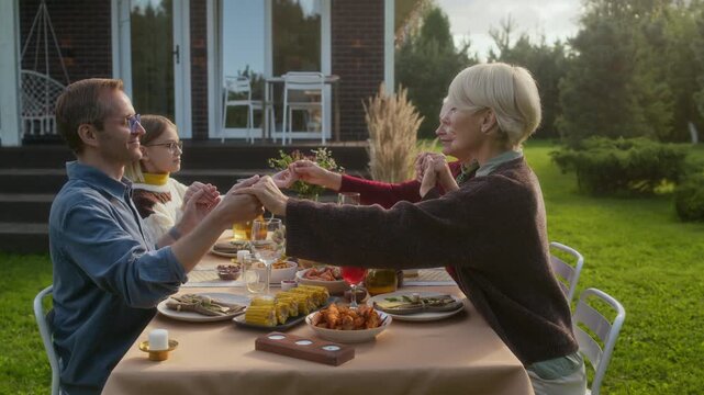Side view shot of multigenerational Caucasian family joining together in prayer before dinner while sitting around dining table during family celebration on warm fall day in cozy backyard outdoors