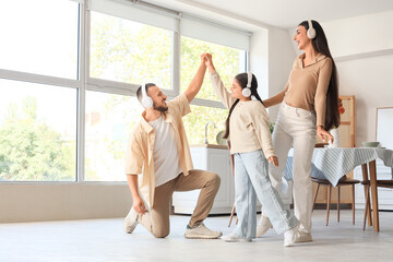 Naklejka premium Happy parents and their little daughter with headphones dancing in kitchen