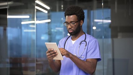 Confident african american doctor in blue coat using digital tablet standing in a modern hospital office. Focused medical worker physician in glasses chats online with patient or works in application - Powered by Adobe