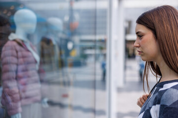 Obraz premium Young stylish woman stands before a boutique window, captivated by elegant clothes and reflections, choosing what to buy with a gentle smile.
