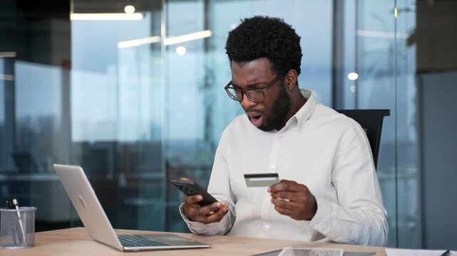 Shocked upset african american businessman checks his credit card on mobile phone in business office. Frustrated male faced fraud, money was stolen from his account. He became a victim of deception