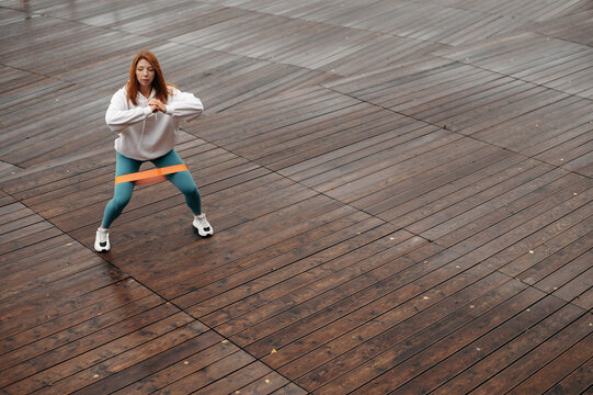 Woman with red hair squats with fitness elastic band on wooden pier