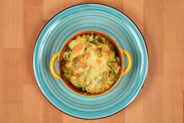 A serving of baked Brussels sprouts and potato with melted cheese in a yellow and brown ceramic cocotte, presented on a turquoise plate with concentric circles