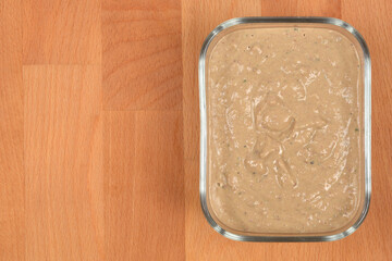 An overhead shot captures a smooth, beige-brown homemade chicken liver pate generously spread inside a rectangular glass container on a light wooden surface, with copy space